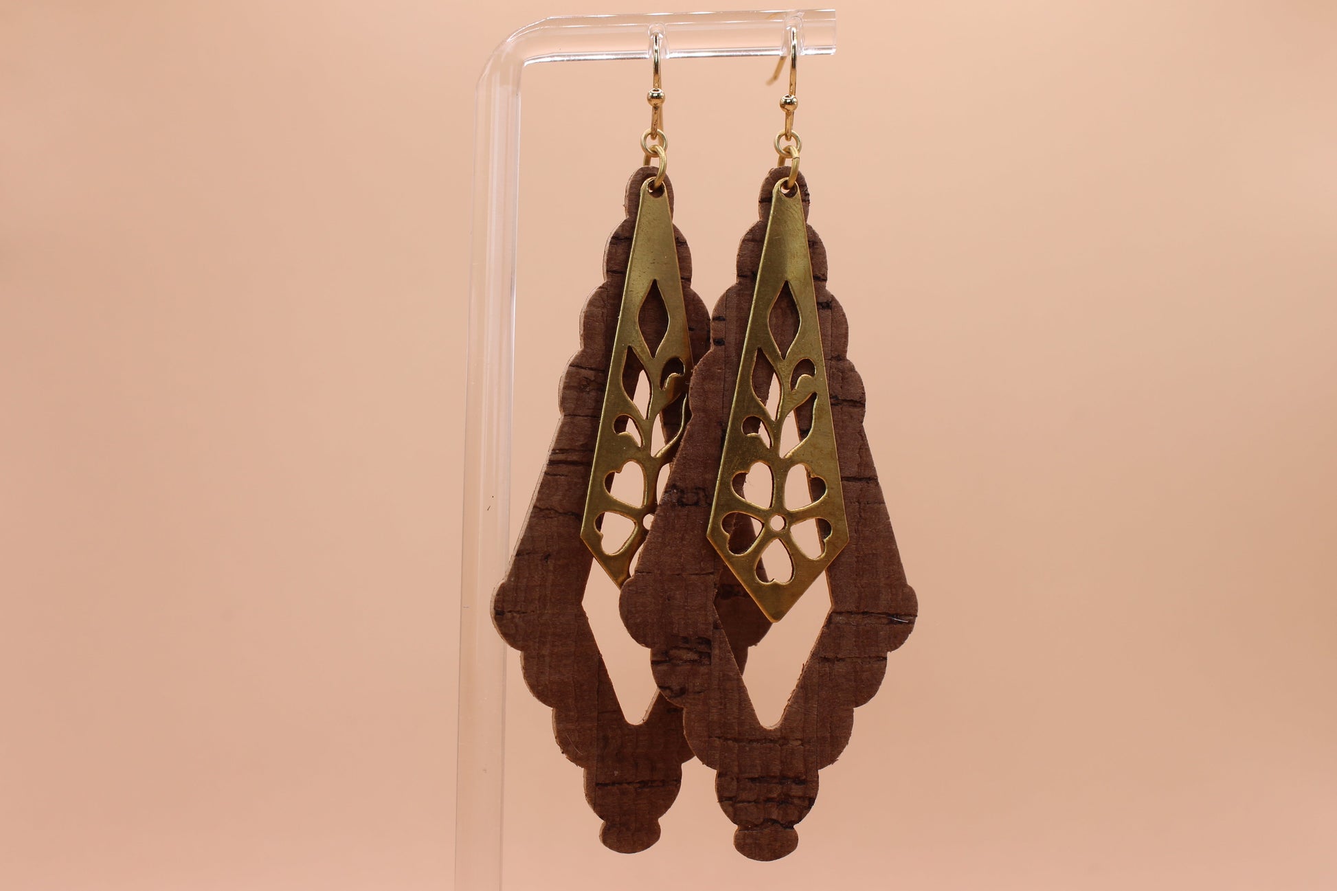 A pair of dangling earrings displayed on a clear acrylic stand against a solid peach background. The earrings feature a gold-toned metal top with a cutout floral pattern over a shaped dark brown cork.