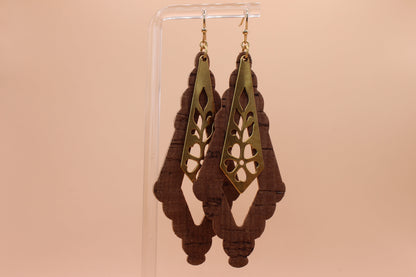 A pair of dangling earrings displayed on a clear acrylic stand against a solid peach background. The earrings feature a gold-toned metal top with a cutout floral pattern over a shaped dark brown cork.