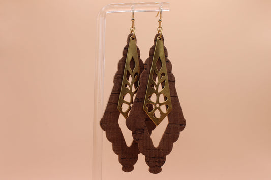 A pair of dangling earrings displayed on a clear acrylic stand against a solid peach background. The earrings feature a gold-toned metal top with a cutout floral pattern over a shaped dark brown cork.
