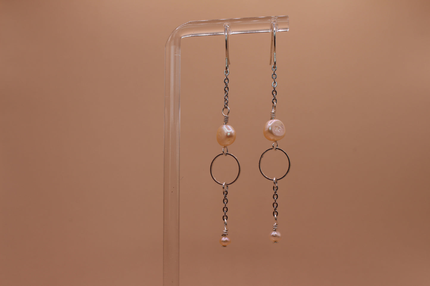 A pair of silver-toned dangle earrings with peach freshwater pearls and circular metal rings, hanging from a clear acrylic display stand against a solid warm tan background.