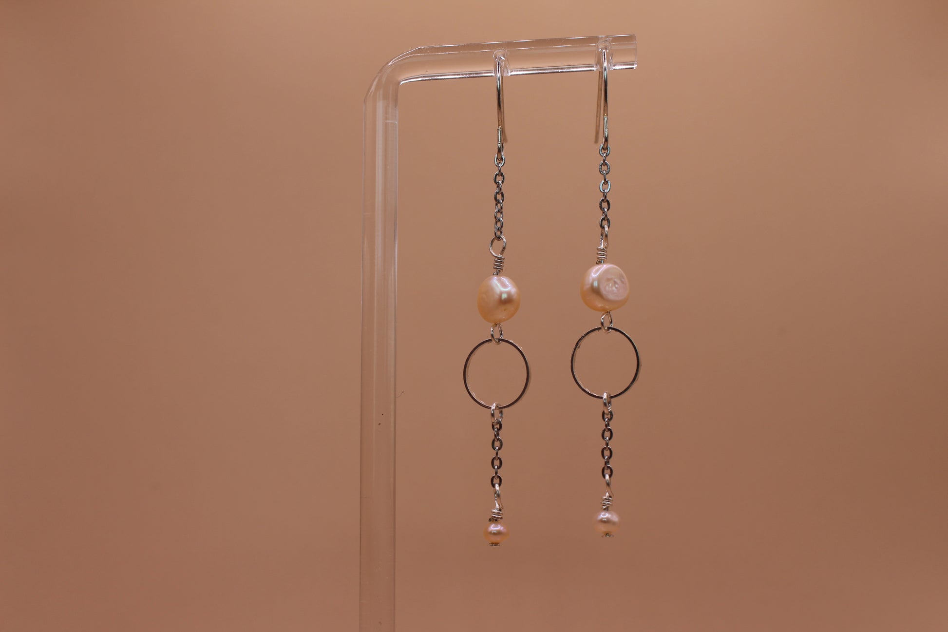 A pair of silver-toned dangle earrings with peach freshwater pearls and circular metal rings, hanging from a clear acrylic display stand against a solid warm tan background.