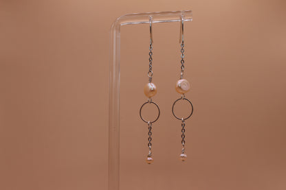 A pair of silver-toned dangle earrings with peach freshwater pearls and circular metal rings, hanging from a clear acrylic display stand against a solid warm tan background.
