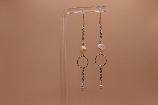 A pair of silver-toned dangle earrings with peach freshwater pearls and circular metal rings, hanging from a clear acrylic display stand against a solid warm tan background.